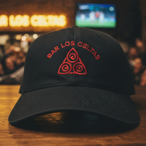 Black baseball cap with red embroidered "Bar Los Celtas" logo and a Celtic-style triangular symbol, placed on a wooden bar counter.